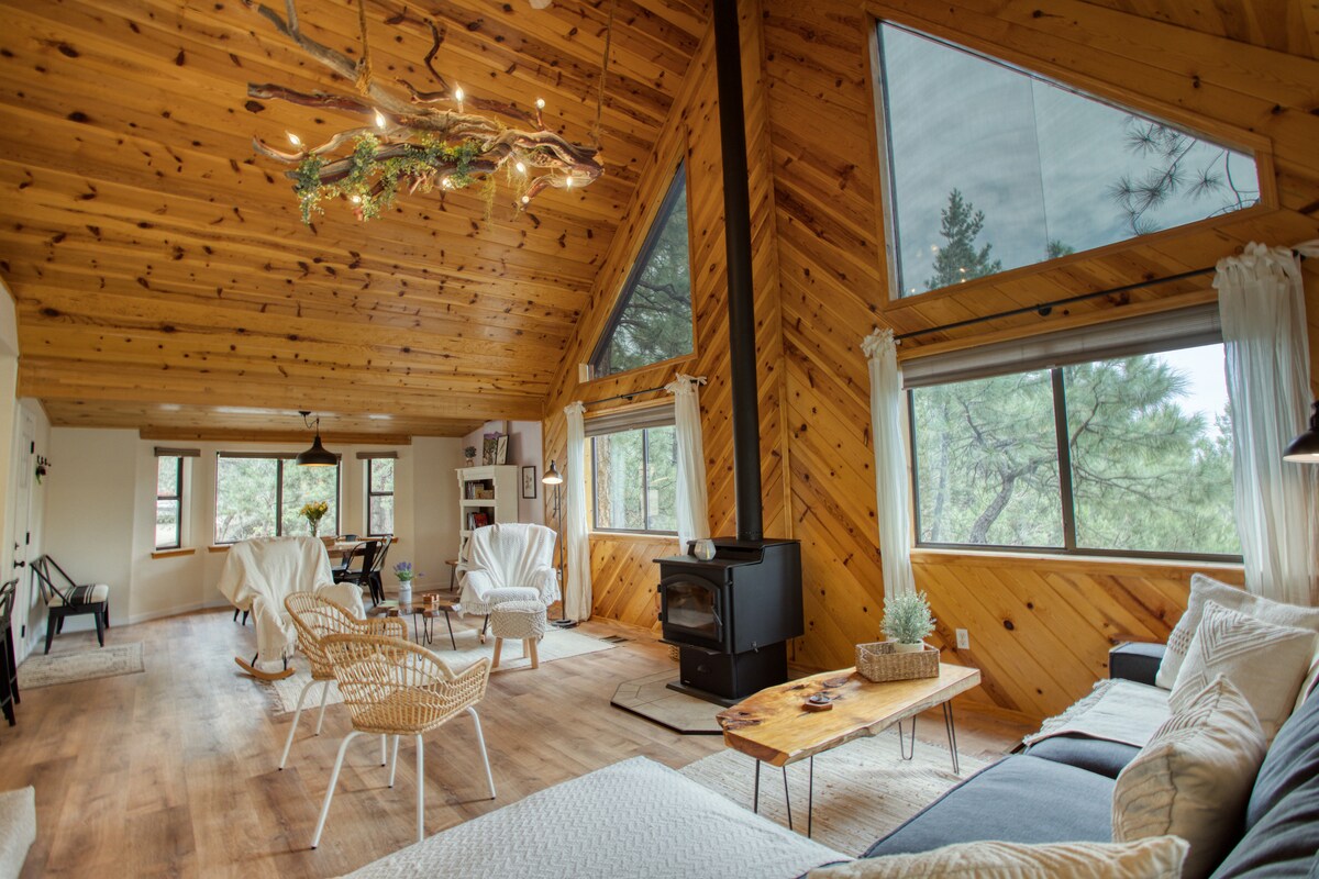 A spacious living area is surrounded by large windows, offering forest views. Natural wood ceilings and walls enhance the cabin's character. A cozy seating arrangement includes two armchairs and a sofa, with a wooden coffee table positioned nearby. A small wood stove adds warmth.