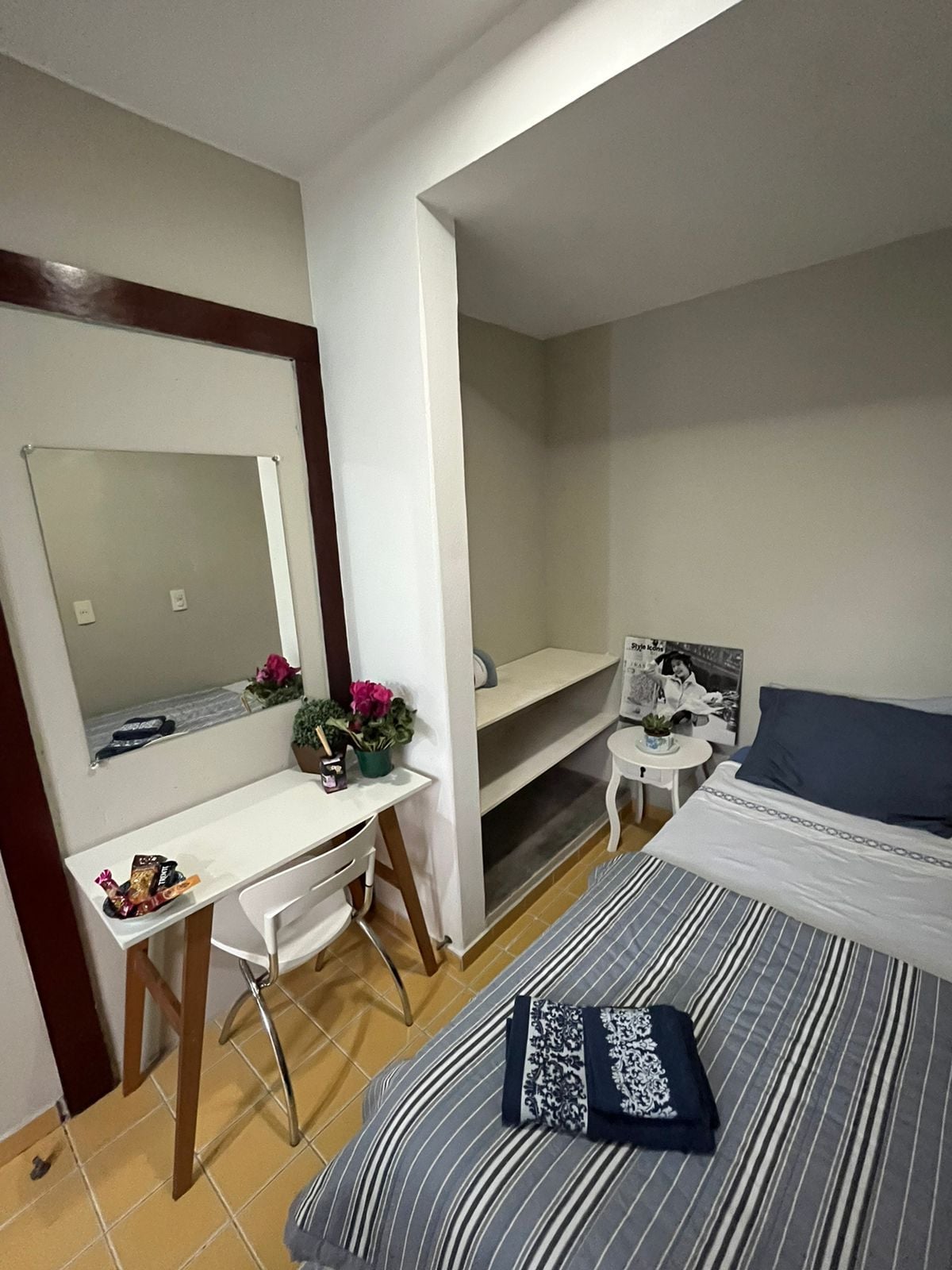 A compact studio space features a spacious bed with gray bedding. A small work desk with a chair sits beside a mirror, complemented by decorative plants. A shelf provides additional storage, with light colors enhancing the overall ambiance.