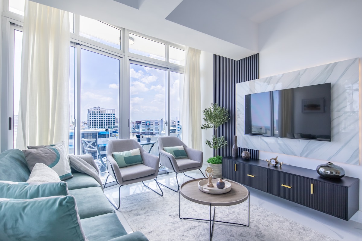 The living room features a modern seating arrangement with a plush sofa and stylish armchairs. A round coffee table sits in the center, complemented by large windows that frame a panoramic view of the city. Soft natural light fills the space, enhancing the contemporary decor.
