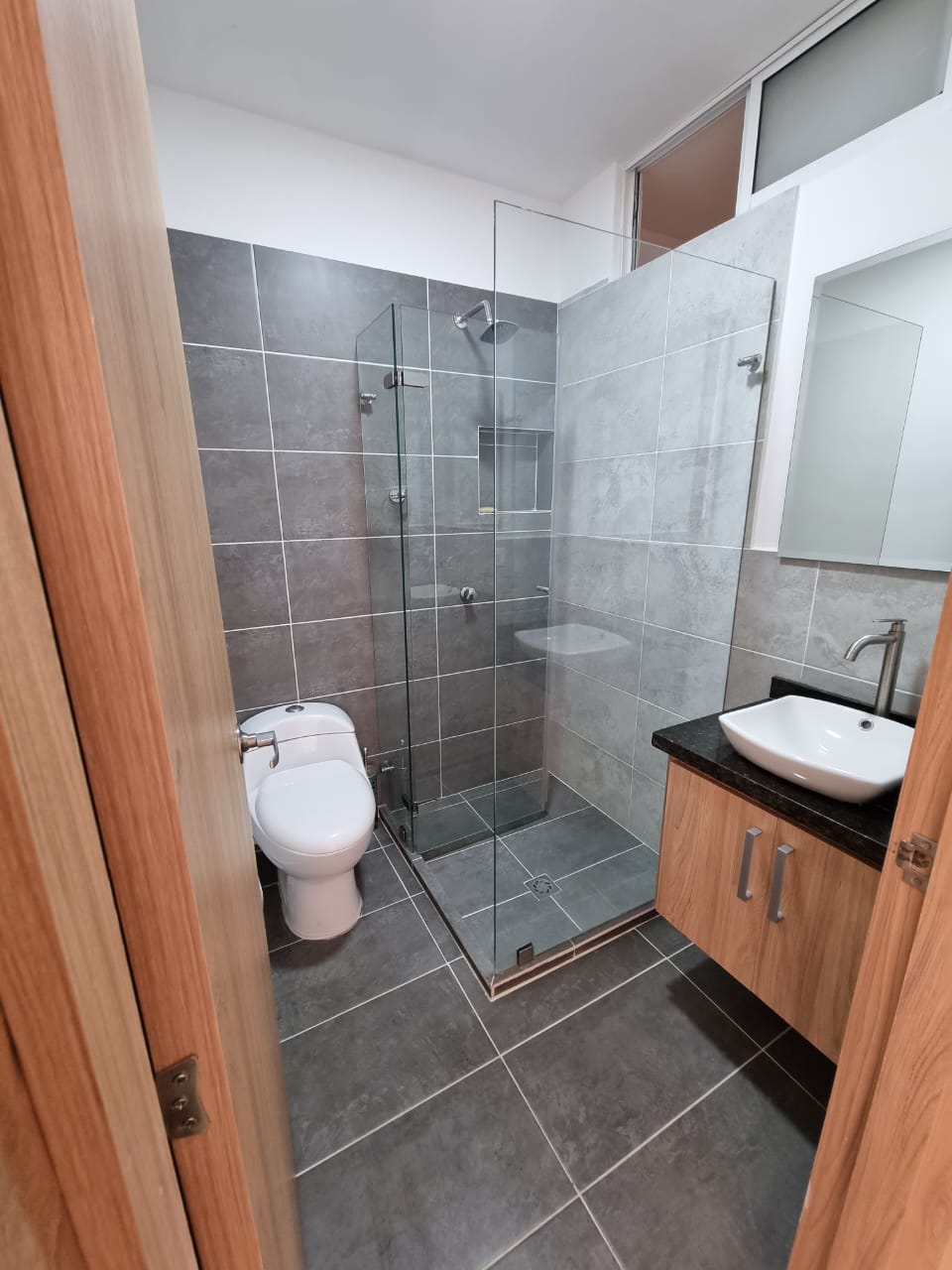A modern bathroom is presented with gray tiled walls and floor. A glass shower enclosure features a rainfall showerhead. The sink area includes a sleek countertop with a mirror above. A white toilet is positioned adjacent to the shower, enhancing the functional layout.