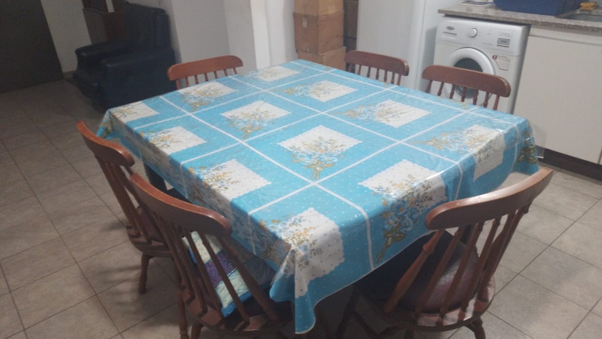A dining area is presented with a large, rectangular table covered by a blue floral tablecloth. Six wooden chairs are arranged around the table, and a washing machine is visible in the background. The space is well-lit, with tiled flooring.