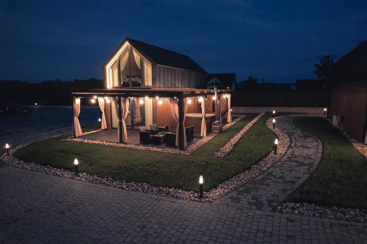 A well-lit cabin exterior is featured at night, surrounded by a landscaped yard. Soft lighting outlines the covered seating area, while a winding stone path leads through the grass. The cabin’s large windows reflect the warm glow, creating a welcoming atmosphere.