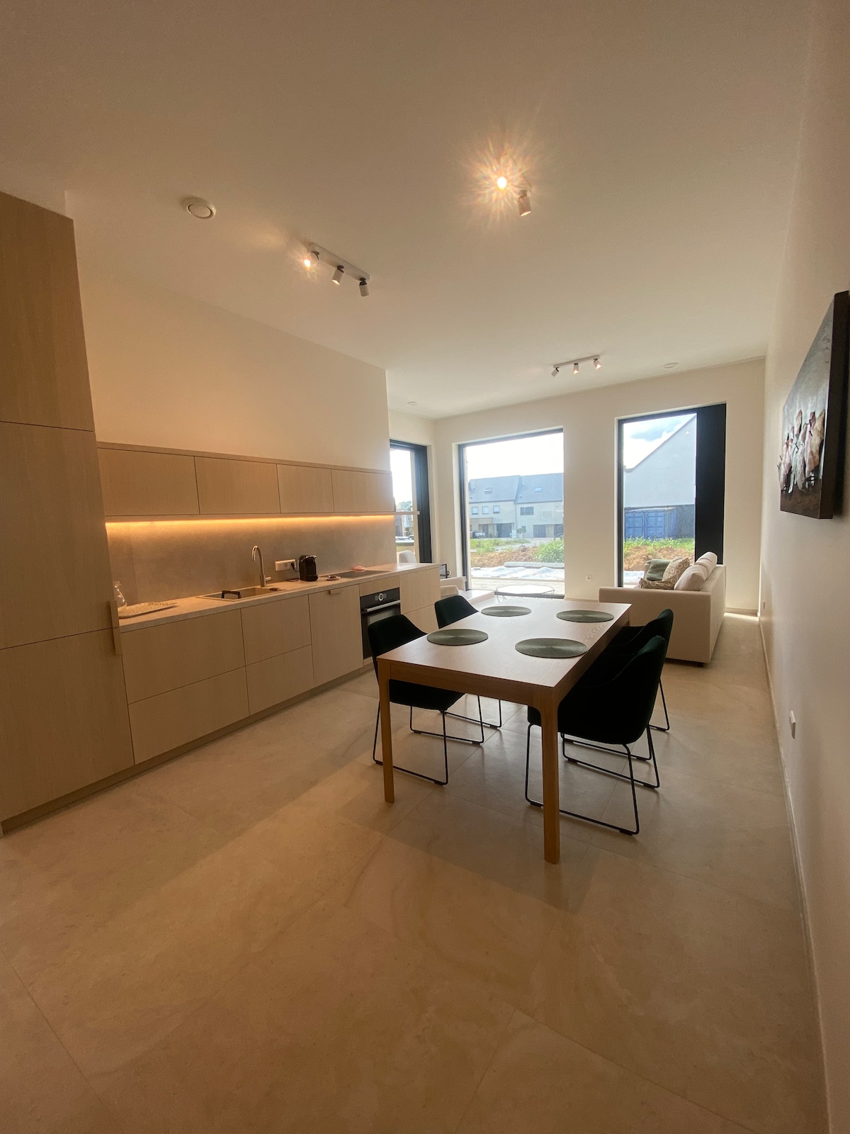 A modern kitchen is featured with wooden cabinetry and sleek countertops. A dining table with four chairs is positioned in the center, complemented by natural light from large windows overlooking the outdoor area. A comfortable sofa is partially visible, enhancing the communal space.