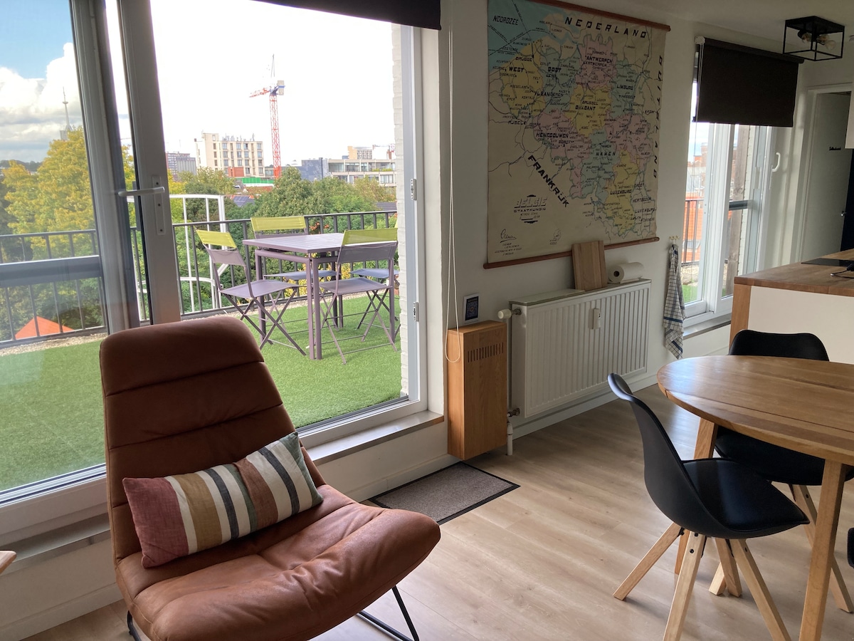 A cozy corner features a brown chair with a striped pillow set beside a round dining table. Large windows allow natural light to fill the room, while a map adorns the wall. A terrace is visible beyond the glass, showcasing outdoor seating.