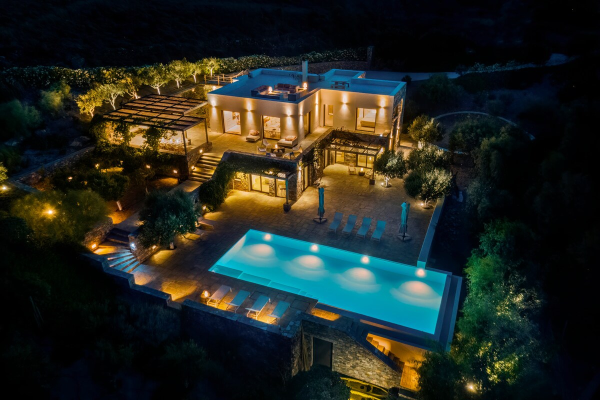 The villa is illuminated at night, showcasing its modern architecture and spacious outdoor areas. A serene swimming pool reflects soft lights, while surrounding terraces reveal inviting seating areas amidst lush greenery. Multiple levels of the villa offer a blend of indoor and outdoor living.