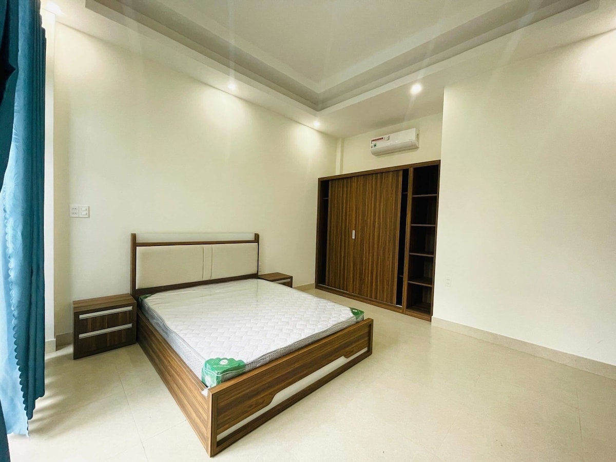 A spacious bedroom is presented, featuring a wooden bed frame without bedding. Flanking the bed are two nightstands. A built-in wardrobe with sliding doors is positioned against one wall. Soft lighting illuminates the room, which has light-colored walls and tiled flooring.