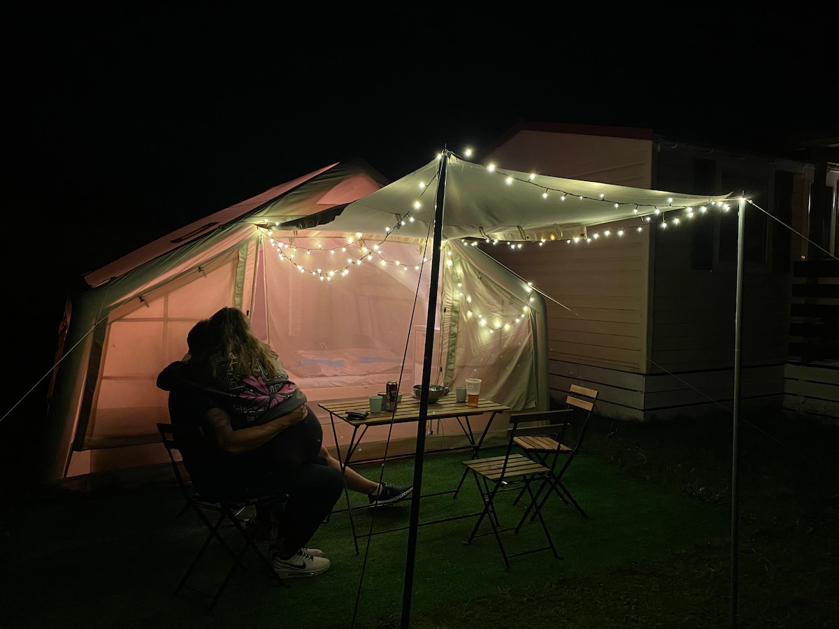 An outdoor glamping area is illuminated by string lights against the night sky. Two chairs are positioned around a table, with a cozy tent visible in the background. The inviting setup is perfect for relaxing and enjoying the outdoors.