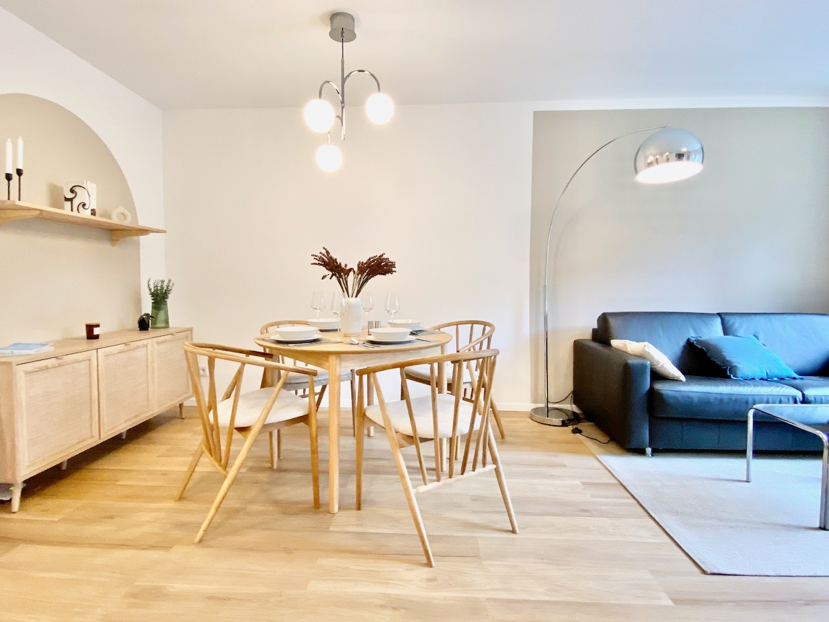 A dining area features a round wooden table surrounded by six light-colored chairs. A modern light fixture hangs above the table, providing soft illumination. A dark sofa, accented with cushions, is positioned nearby, and a stylish floor lamp adds additional light to the space.