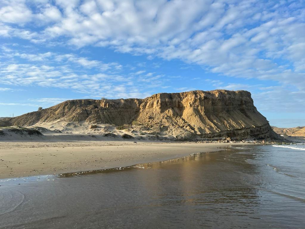 A sandy beach extends towards a prominent rock formation, characterized by layered cliffs under a sky filled with scattered clouds. The serene water reflects the landscape, creating a calm and balanced view of nature.