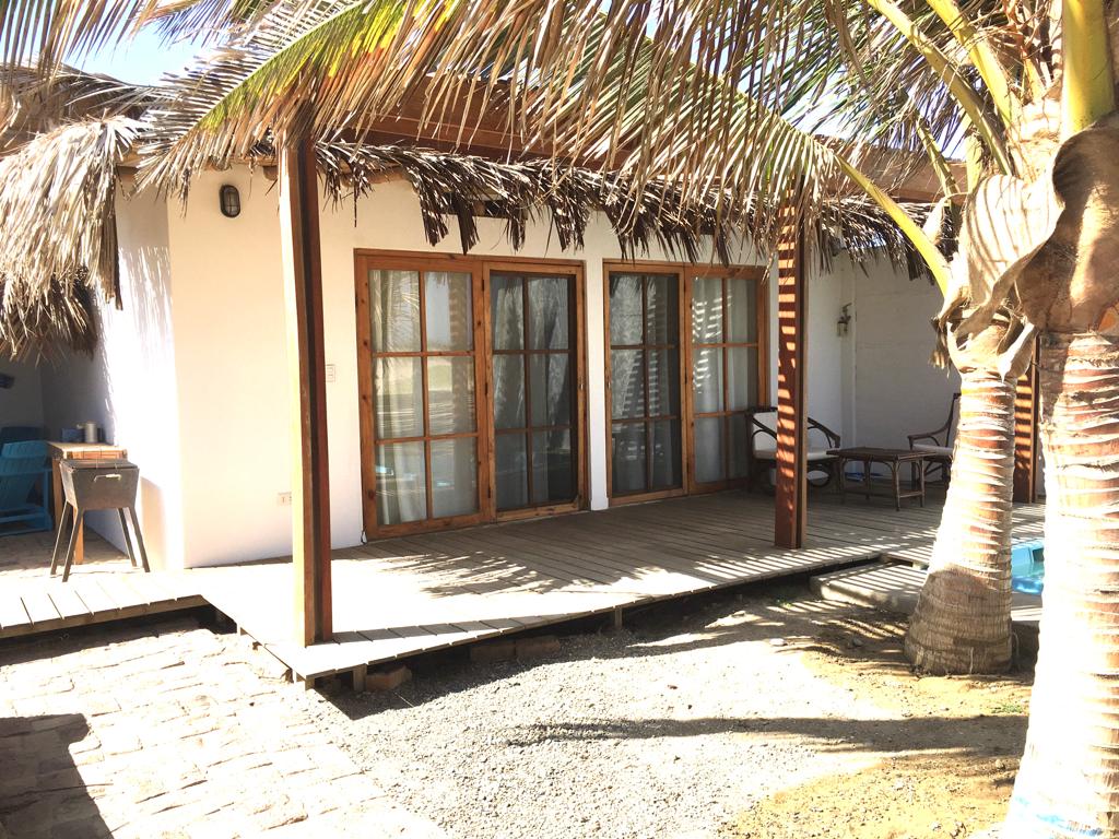 A wooden deck leads to a bungalow entrance, framed by tall palm trees. Glass doors provide access to the interior, flanked by a pair of comfortable outdoor chairs. The natural surroundings enhance a serene atmosphere, while the shaded area invites relaxation.