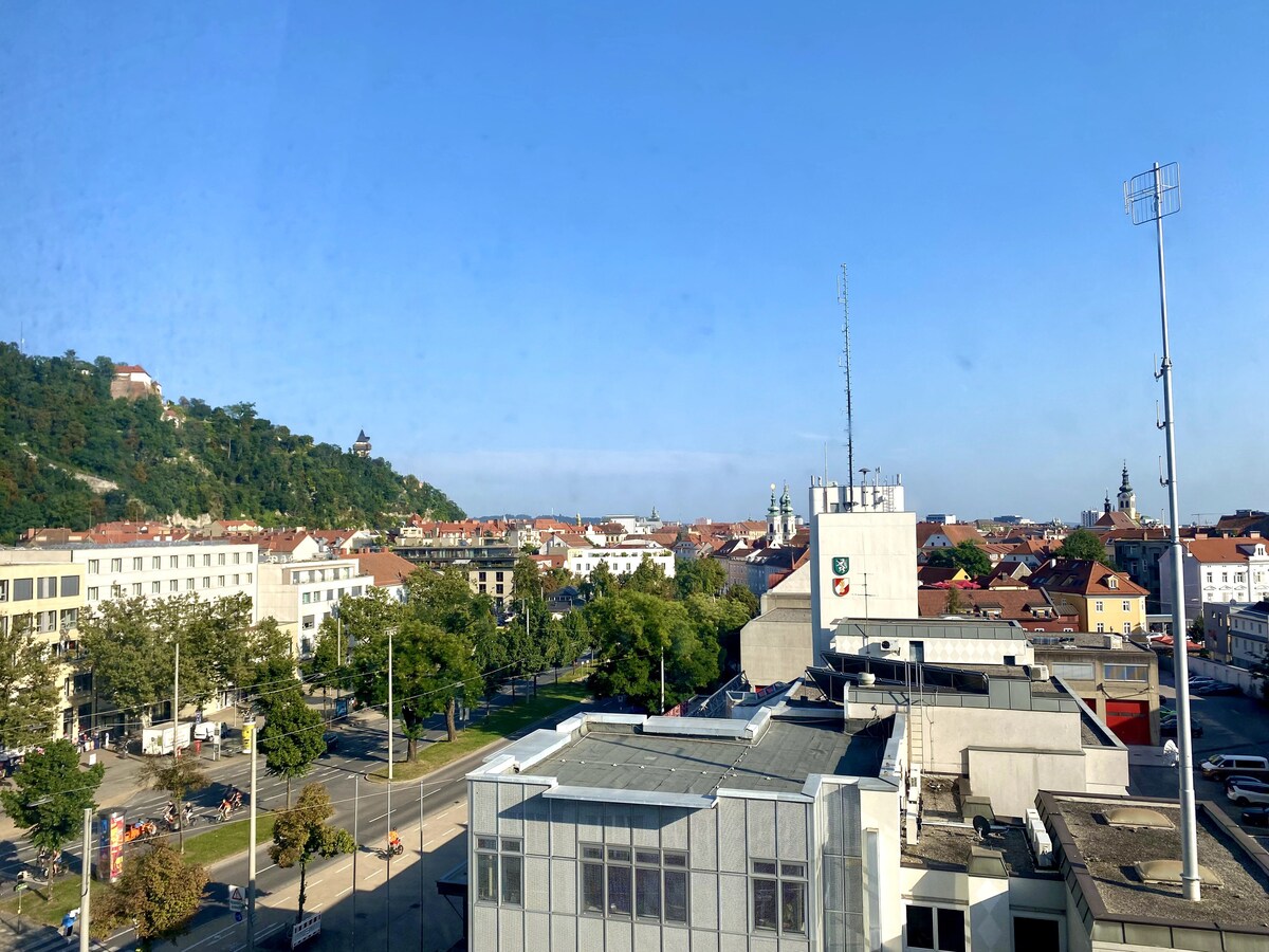 A panoramic view showcases the skyline of Graz, featuring varied architectural styles of buildings and greenery. The Schlossberg hill rises prominently in the distance, while bright blue skies provide a clear backdrop. The image captures the lively atmosphere of the city from an elevated perspective.