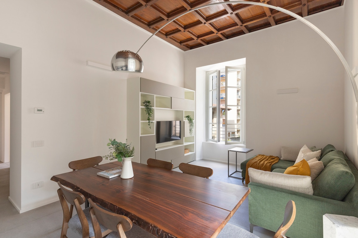 A spacious living area features a wooden dining table with matching chairs and a green sofa. A modern shelving unit with plants displays a television. Natural light floods through the large window, highlighting the intricately designed wooden ceiling.