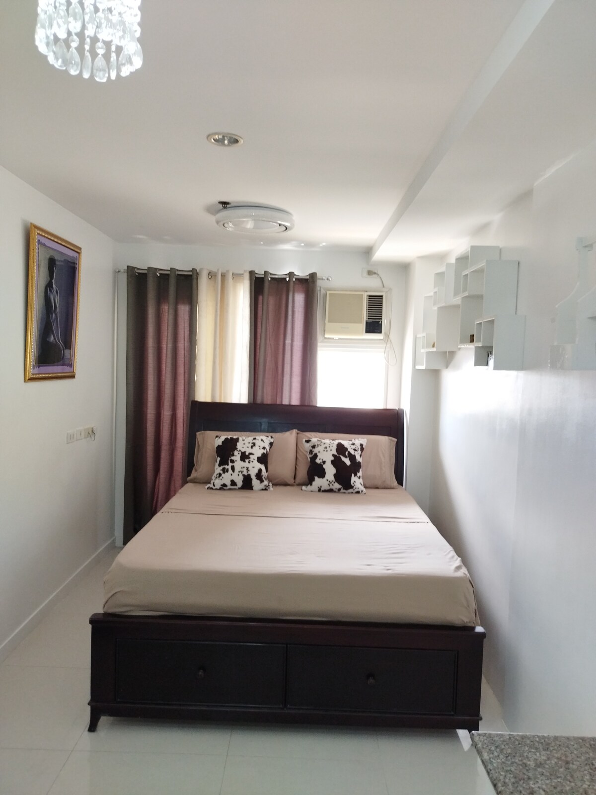 A cozy sleeping area features a spacious bed with neutral bedding and decorative pillows. Softly draped curtains in subtle tones frame a window. A wall-mounted air conditioning unit and minimalist shelving enhance the functionality of the room, while a framed artwork adds a decorative touch.