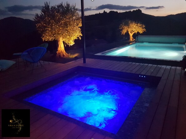 A luxurious spa with illuminated blue water sits next to a spacious pool, both surrounded by wooden decking. Two olive trees are softly lit by garden lights, adding an elegant touch to the serene outdoor setting at dusk.