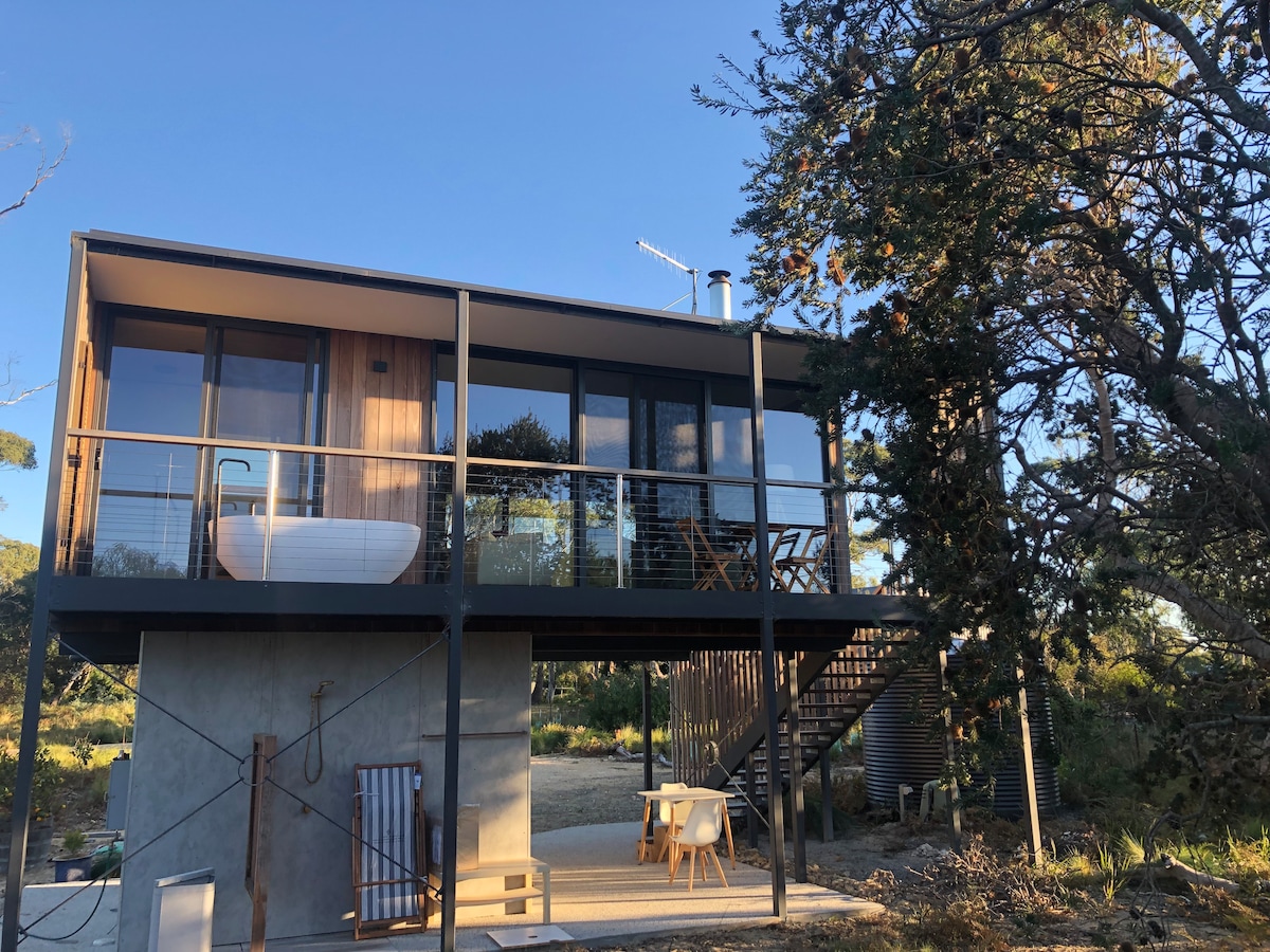 The studio's modern exterior showcases large windows and a private deck with a freestanding outdoor bath. A staircase leads up to the entrance, surrounded by natural landscaping. A small outdoor seating area is visible on the ground level, offering a welcoming space for relaxation.
