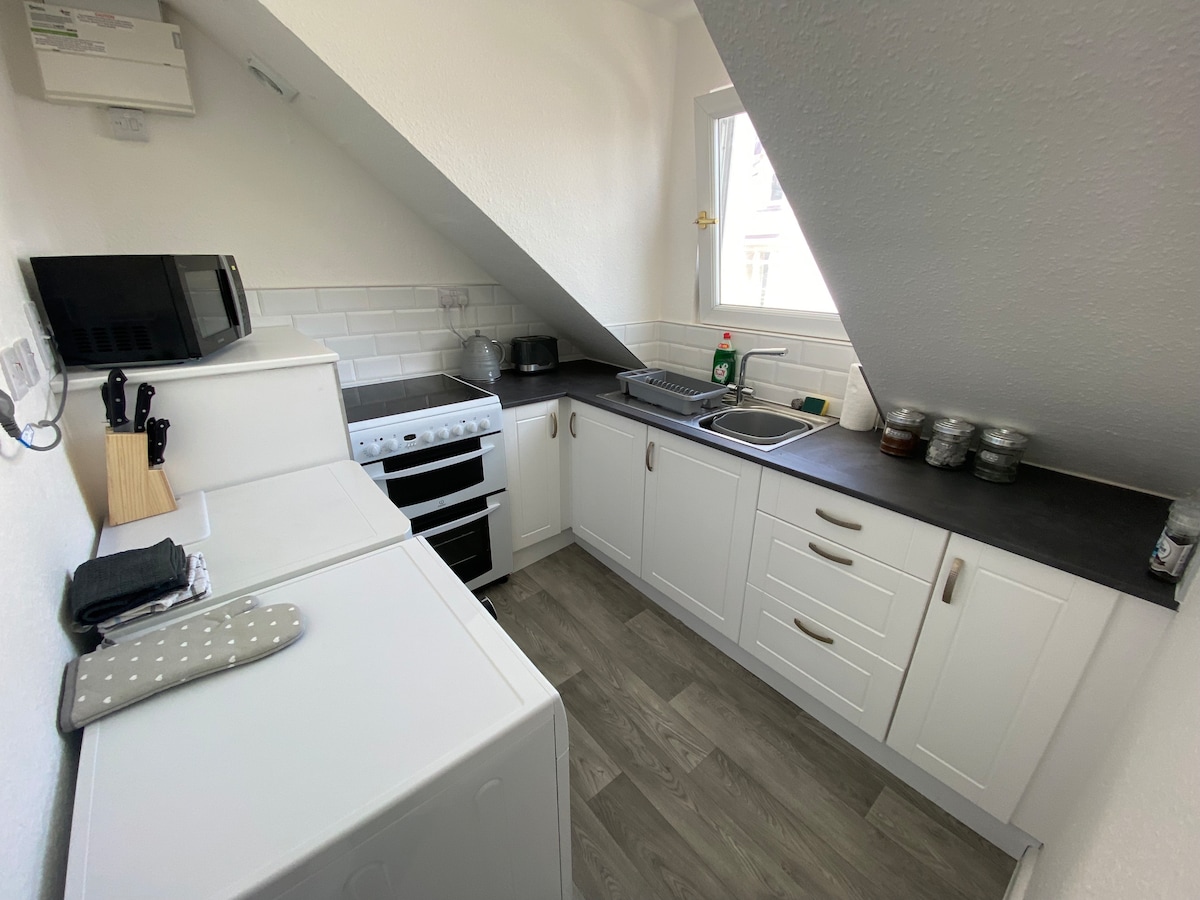 A compact kitchen features modern white cabinetry and a dark countertop, with essential appliances including a microwave and a stove. A sink is positioned under a window, allowing natural light to brighten the space. The floor is covered in light-colored vinyl planks.