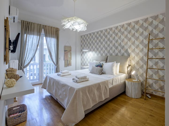 Bright and elegant bedroom featuring a plush king-sized bed, stylish décor, and a smart TV. Enjoy natural light streaming through large windows with charming curtains, complemented by a cosy balcony view. Perfect for a serene and relaxing stay.