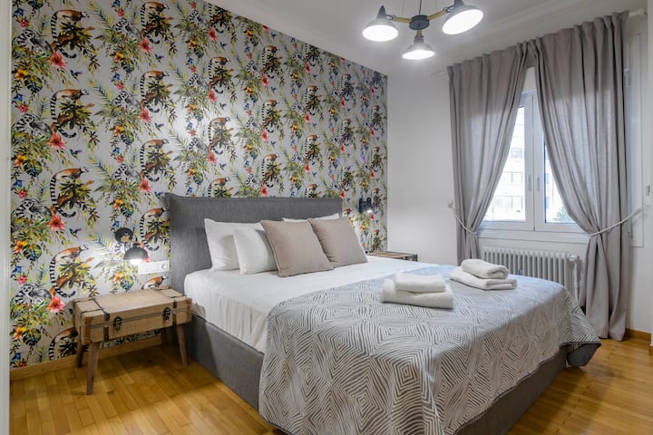 A bright and inviting bedroom featuring a luxurious king-sized bed, elegant patterned wallpaper, wooden accents, and soft lighting. Large windows with flowing curtains create a serene atmosphere, perfect for relaxation.