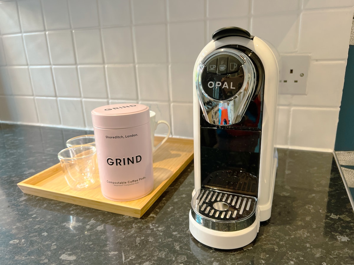 A sleek coffee machine is positioned on a dark countertop, accompanied by a pink container labeled 'GRIND' holding compostable coffee pods. Two clear glasses sit on a wooden tray beside the machine. White tiled walls provide a clean backdrop.