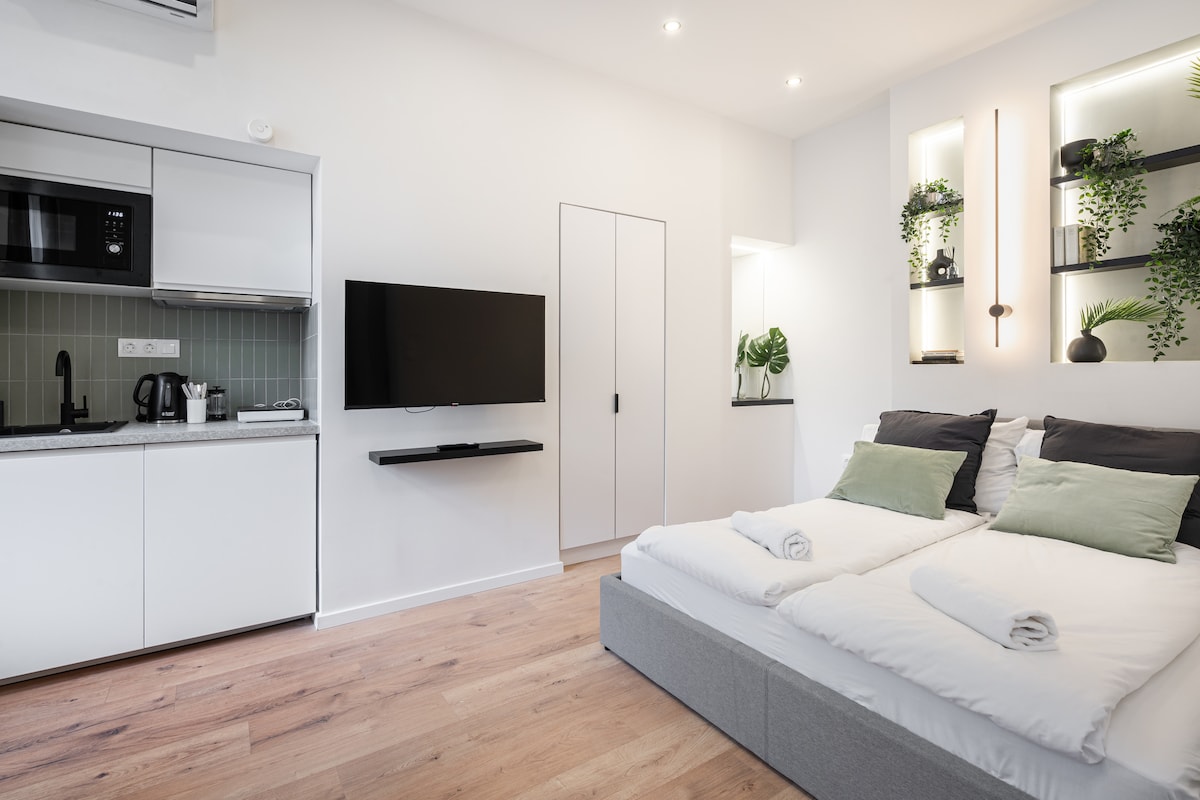 A modern studio apartment features a spacious bed with white linens and rolled towels. A flat-screen TV is mounted on the wall, while a kitchenette is visible, equipped with cooking appliances. Green plants adorn shelves, adding a touch of nature to the clean, minimalist design.