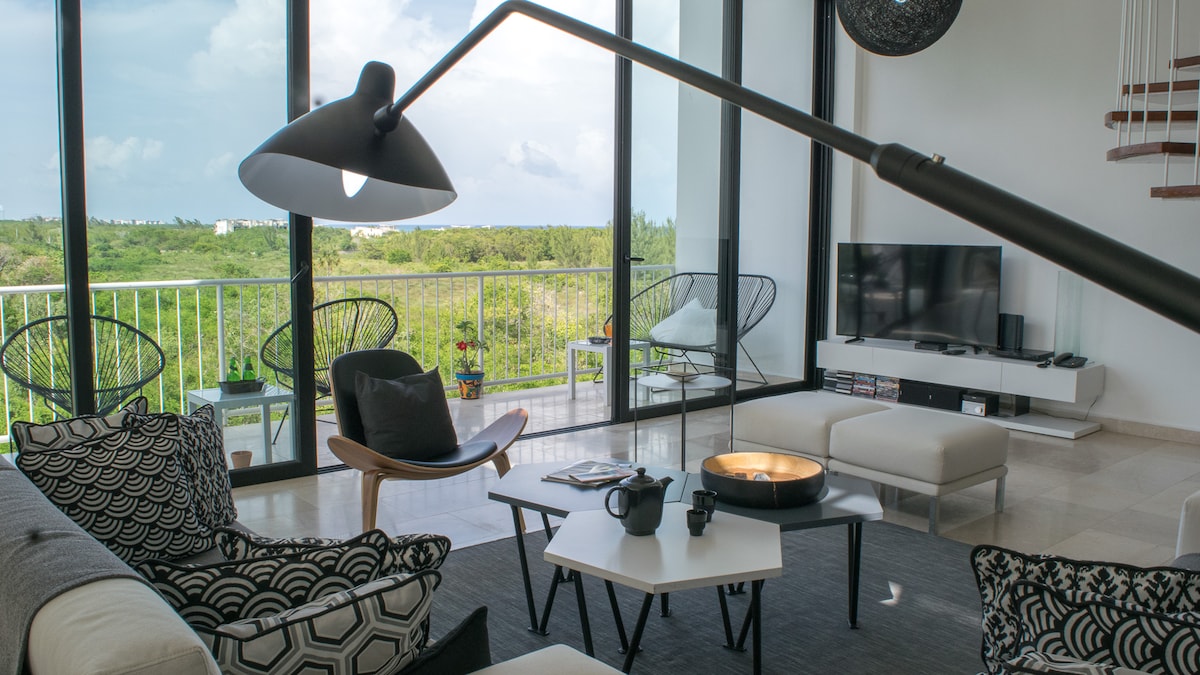 A spacious living area features large glass doors revealing a green landscape. A modern coffee table is centered in front of a low couch, accompanied by stylish armchairs. A sleek floor lamp provides soft lighting, while a television is positioned on a light display unit.