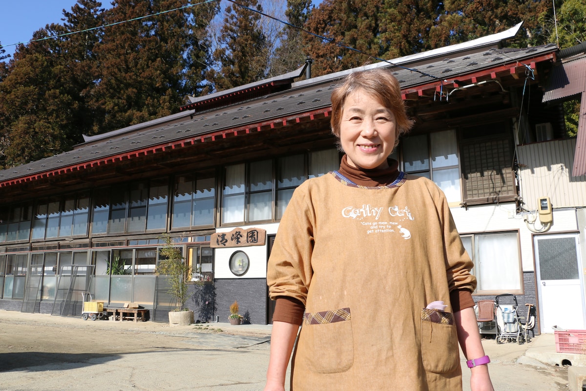 The exterior of a large traditional house is visible, characterized by its wooden structure and large overhanging roof. The building's entrance is adorned with signage, while the surrounding landscape features trees typical of the area, adding to the serene atmosphere.