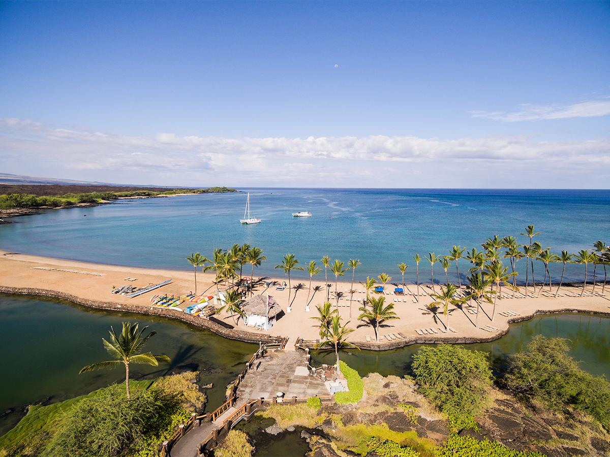 An aerial view captures a sandy beach bordered by palm trees, leading to calm blue waters. A few sailing boats are visible on the horizon. In the foreground, a pathway curves alongside a lagoon, enhancing the natural landscape.
