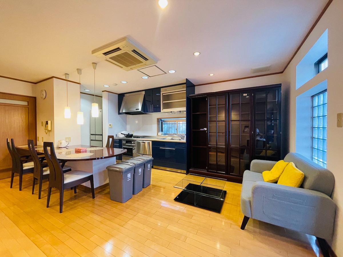An open living space is presented featuring a dining table with chairs, a modern kitchen with dark cabinetry, and a comfortable sofa with cushions. Light wood flooring enhances the bright atmosphere, while a glass-top table adds a functional touch. Natural light enters through the windows.