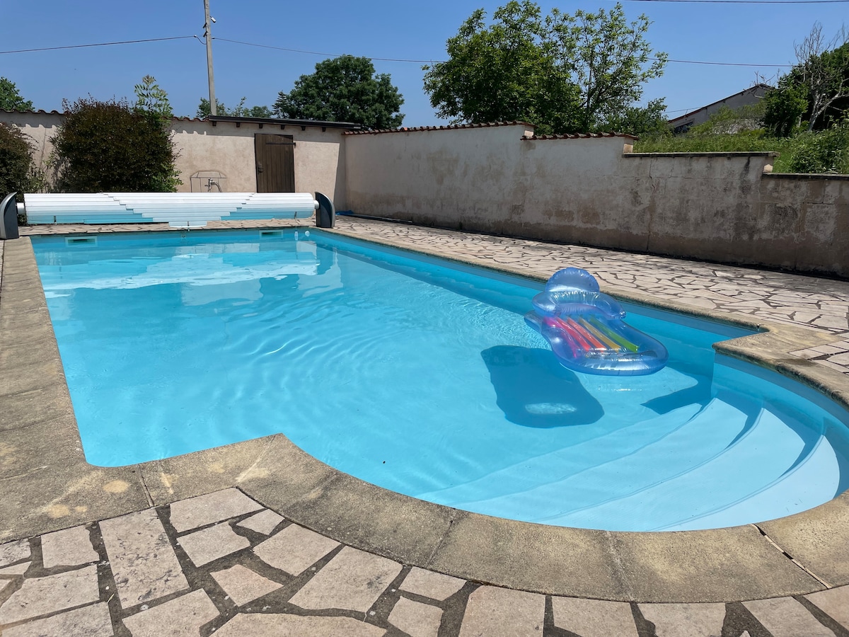 A rectangular swimming pool is situated in a sunlit outdoor area, featuring light blue water and a colorful floating lounge. Surrounding the pool, a textured stone patio provides ample space for relaxation, bordered by a low wall and greenery in the distance.