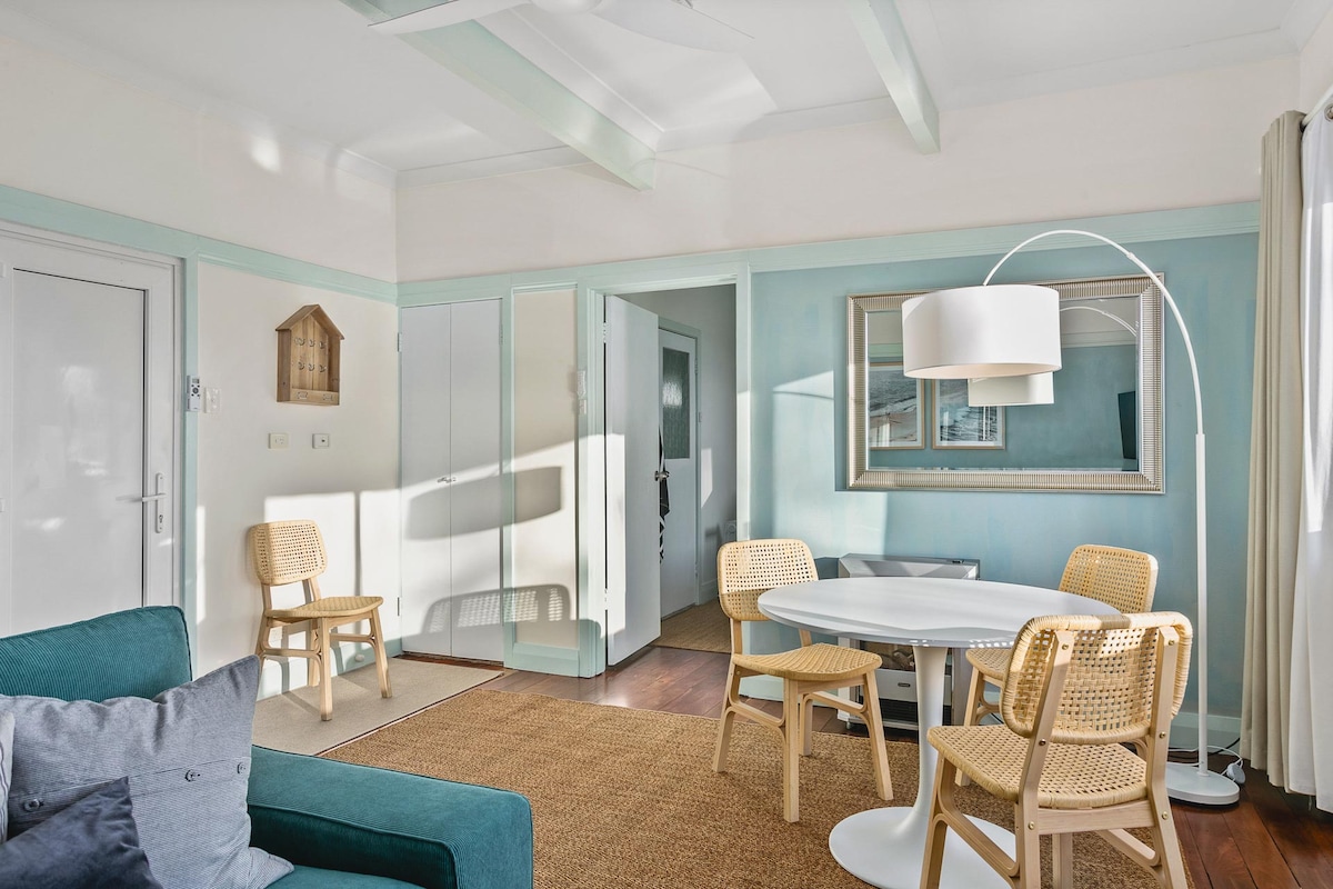 A bright and inviting living space features large windows allowing natural light to fill the room. A round dining table with four chairs is complemented by a light fixture, while a cozy green couch and neutral rug complete the decor. Walls are painted in soft, calming colors.