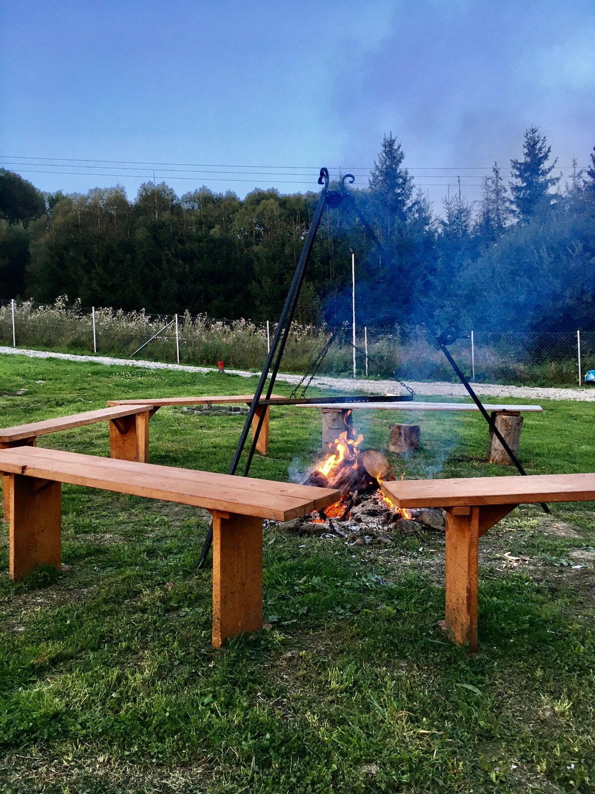 A fire pit surrounded by wooden benches is shown in a grassy area. Smoke rises from the fire, creating a cozy atmosphere. A natural backdrop of trees is visible in the distance, adding to the outdoor setting.