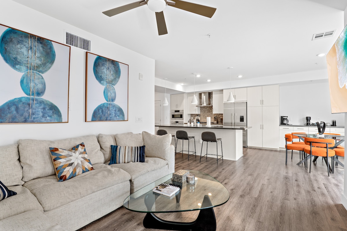 An open-concept living area is highlighted by a spacious light-colored sectional sofa, accompanied by a round glass coffee table. A modern kitchen with sleek appliances and a large island is visible in the background, while a dining area with vibrant orange chairs is positioned nearby.