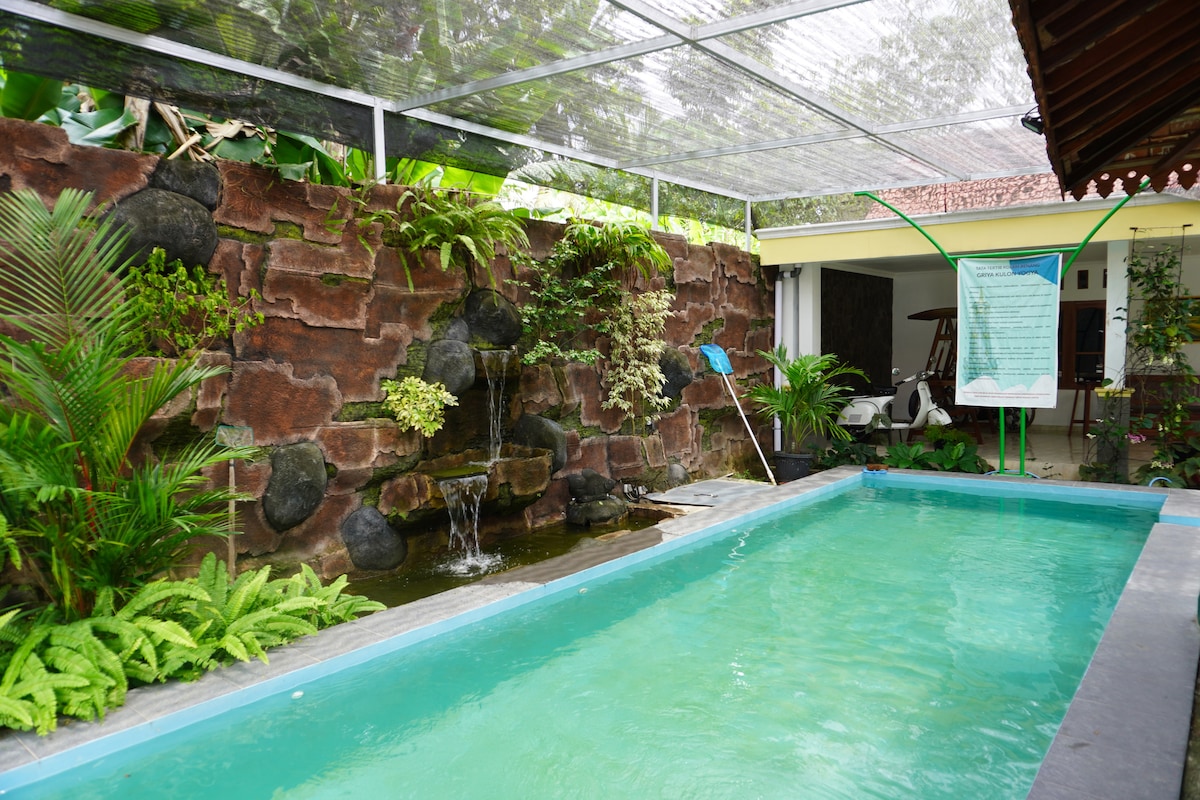 A serene swimming pool is surrounded by lush greenery, featuring a rock wall and a gentle water feature. Ample natural light filters through the overhead cover, creating a calming atmosphere. The area is designed for relaxation, enhancing the overall tranquil setting.