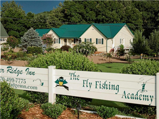 A charming inn is depicted, featuring a green metal roof and light-colored siding. Surrounding gardens are landscaped with various plants and trees. A welcoming sign indicates the presence of a fishing academy, enhancing the serene, outdoor atmosphere.
