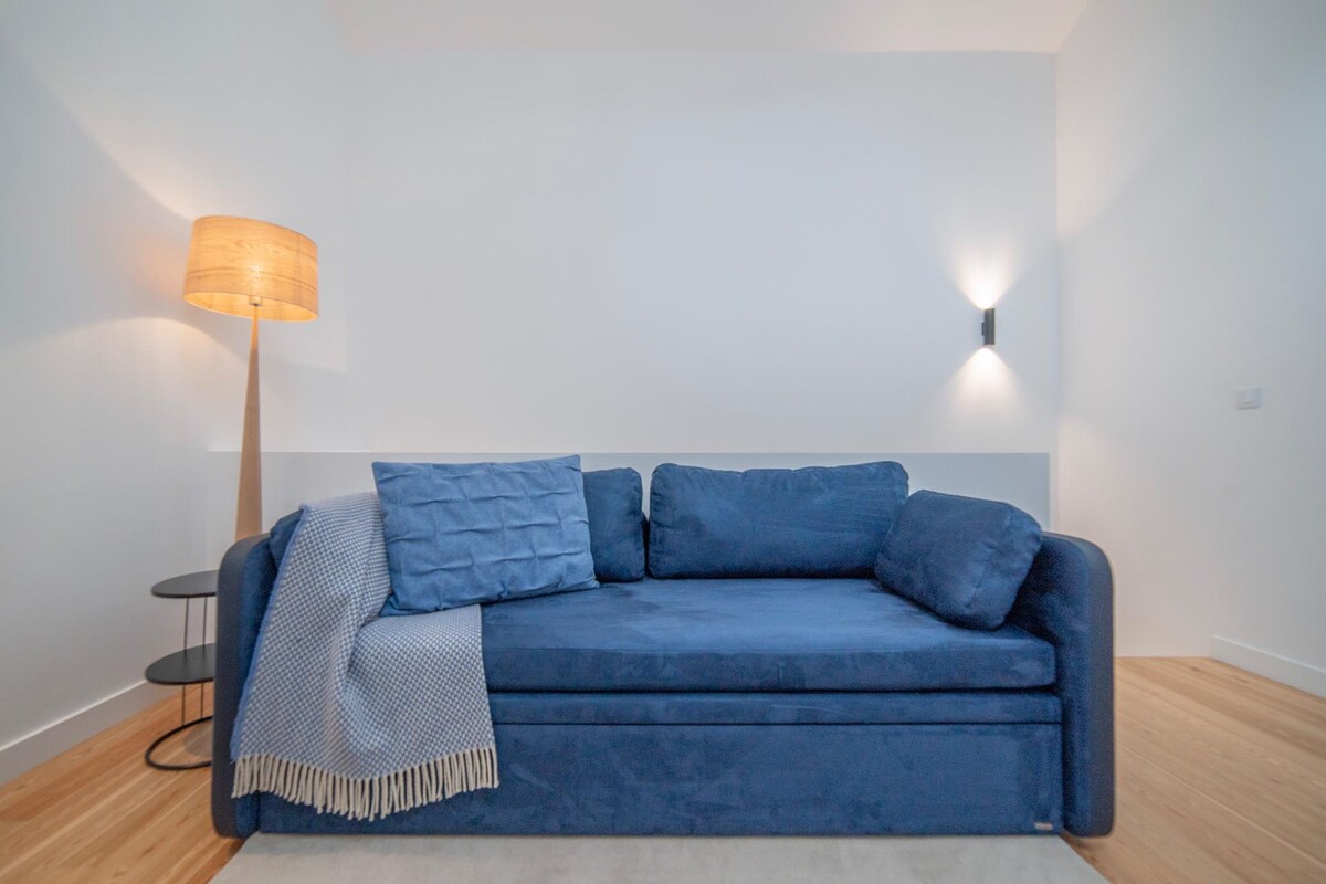 A modern, inviting couch in deep blue features a textured throw blanket and accent pillows. Soft lighting is provided by a nearby floor lamp, creating a warm ambiance. A small side table complements the seating area, enhancing its functional design.