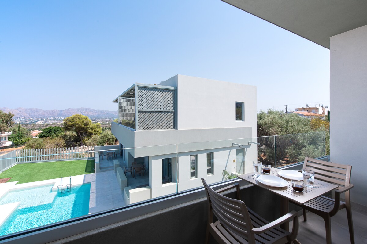 A spacious balcony is shown, featuring a table set with two glasses and plates, overlooking a modern residence with a pool. The structure showcases clean lines and large windows, surrounded by greenery under a clear blue sky.