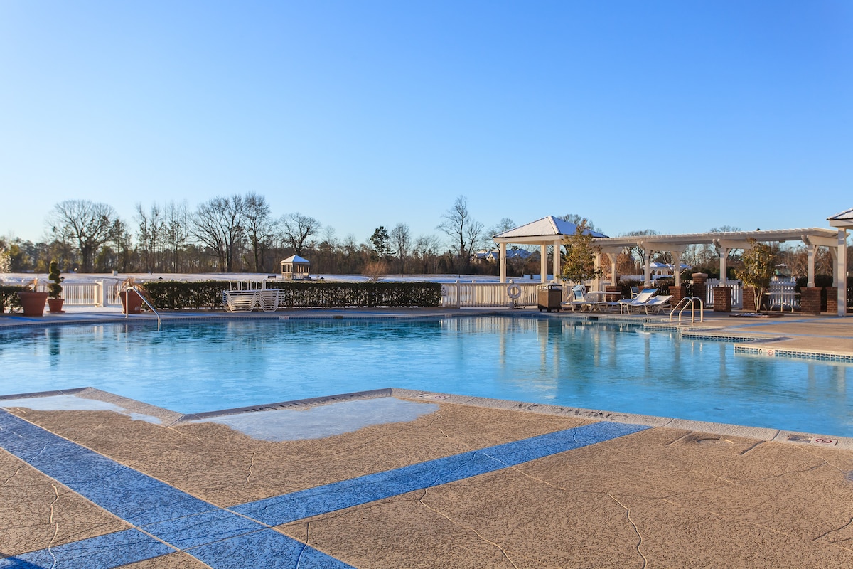 A large outdoor swimming pool invites relaxation, with clear blue water reflecting the sky. Surrounding the pool are well-maintained lounge areas and landscaped greenery. Sunlight enhances the calm atmosphere, and nearby structures provide shaded seating options.