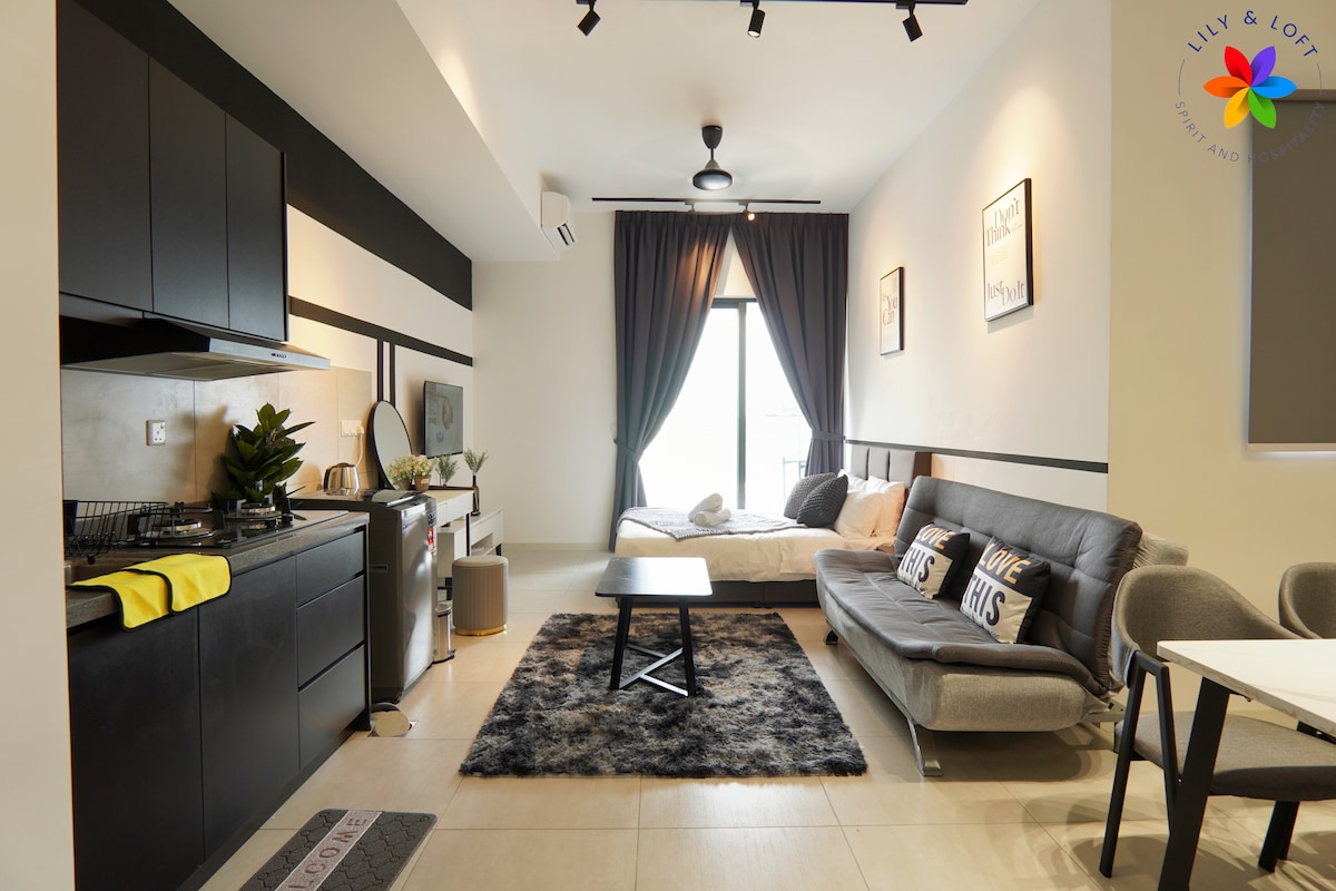 A modern studio suite features a comfortable gray sofa, a dining table with chairs, and a spacious bed bathed in natural light from large windows. The kitchenette includes essential appliances. A plush area rug adds a cozy touch to the space.