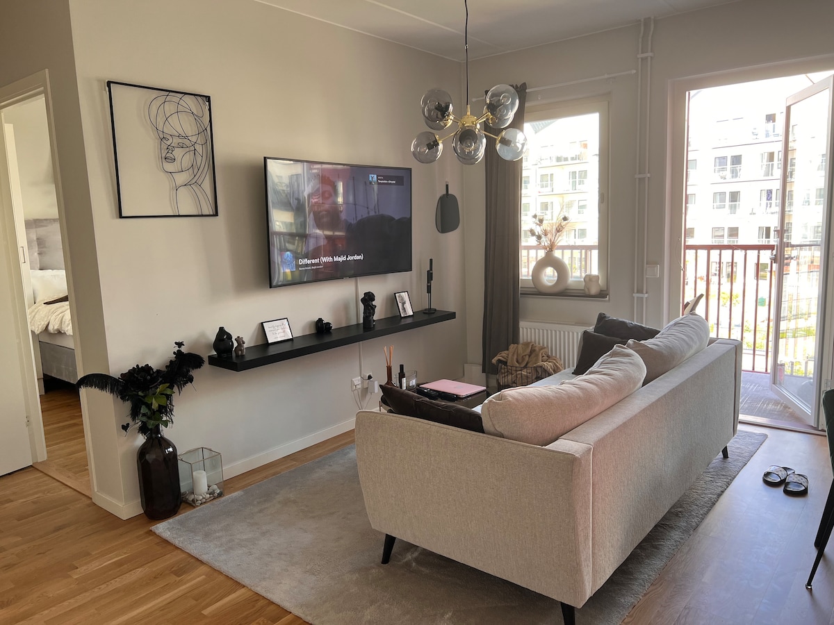 A light-filled living area features a plush sofa facing a wall-mounted television. A sleek shelf holds decorative items and plants. Large windows provide access to a balcony, enhancing the natural light, while a minimalist light fixture adds a modern touch to the space.