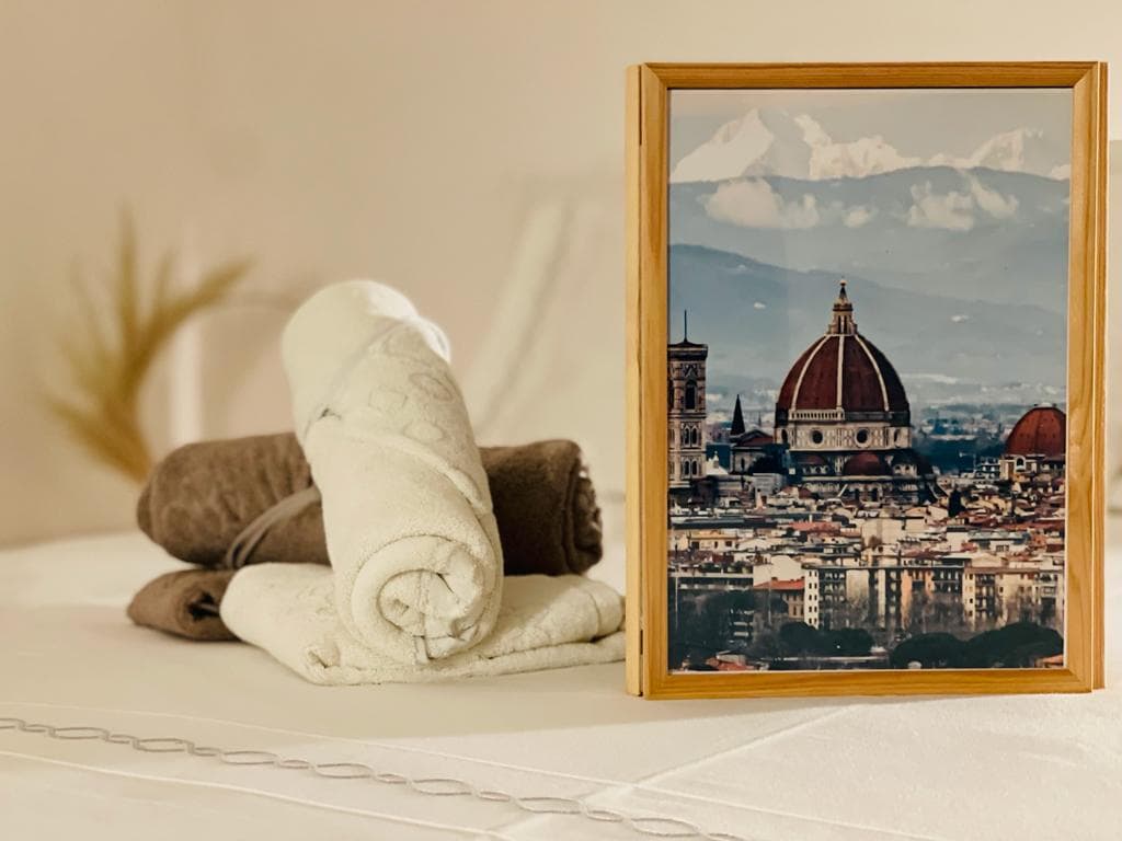 A soft-focus arrangement features neatly folded towels in neutral tones alongside a framed photograph of Florence's skyline. The photograph highlights the distinctive dome of the cathedral, while subtle decorative elements enhance the serene setting.