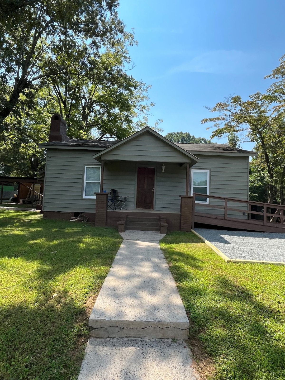 A charming house features gray siding and a sloped roof. A concrete walkway leads to an entrance with steps flanked by brick columns. A small porch sits at the front, containing a couple of chairs. Lush greenery surrounds the property, adding to its serene setting.
