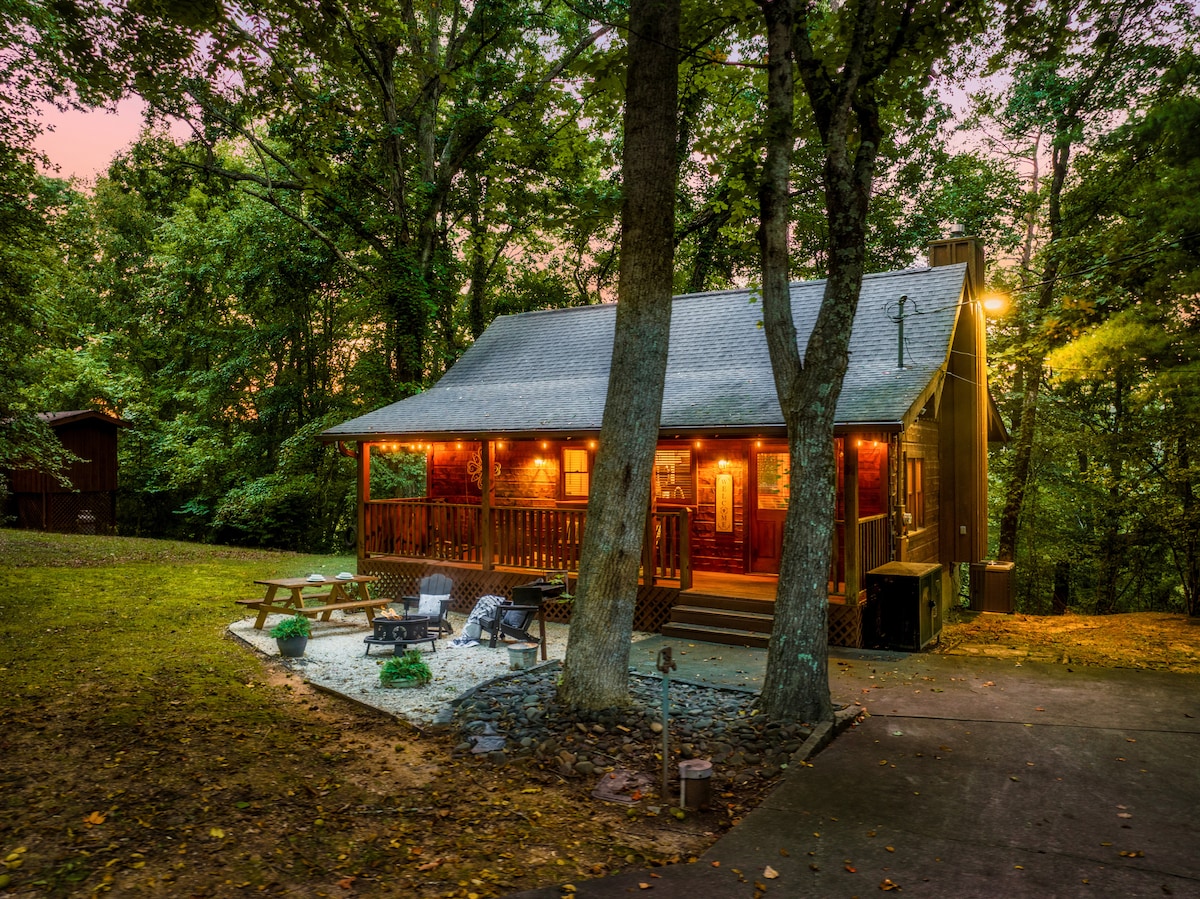 The cabin is nestled among tall trees, featuring a covered porch with warm outdoor lighting. A stone fire pit is situated in the yard, surrounded by seating options, including picnic tables and Adirondack chairs. The natural surroundings create a serene atmosphere.