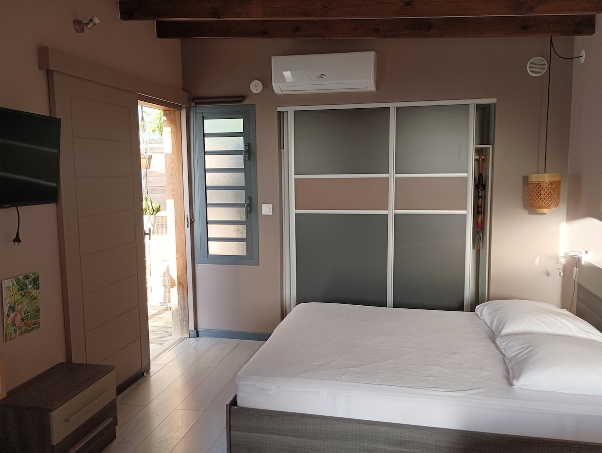 The bedroom includes a spacious bed with white bedding and pillows. Natural light streams through the partially open entrance door, highlighting a neutral color palette. A wall-mounted television is visible, alongside a modern sliding door closet and a small bedside lamp.
