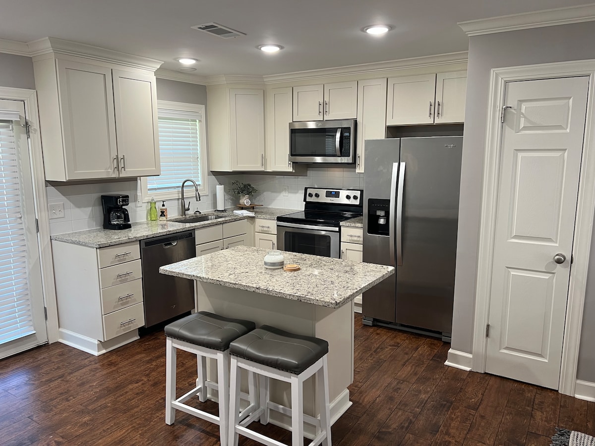A modern kitchen is displayed with light-colored cabinetry and granite countertops. Stainless steel appliances include a refrigerator, oven, and microwave. A central island with seating for two is shown, complemented by a window allowing natural light to brighten the space.
