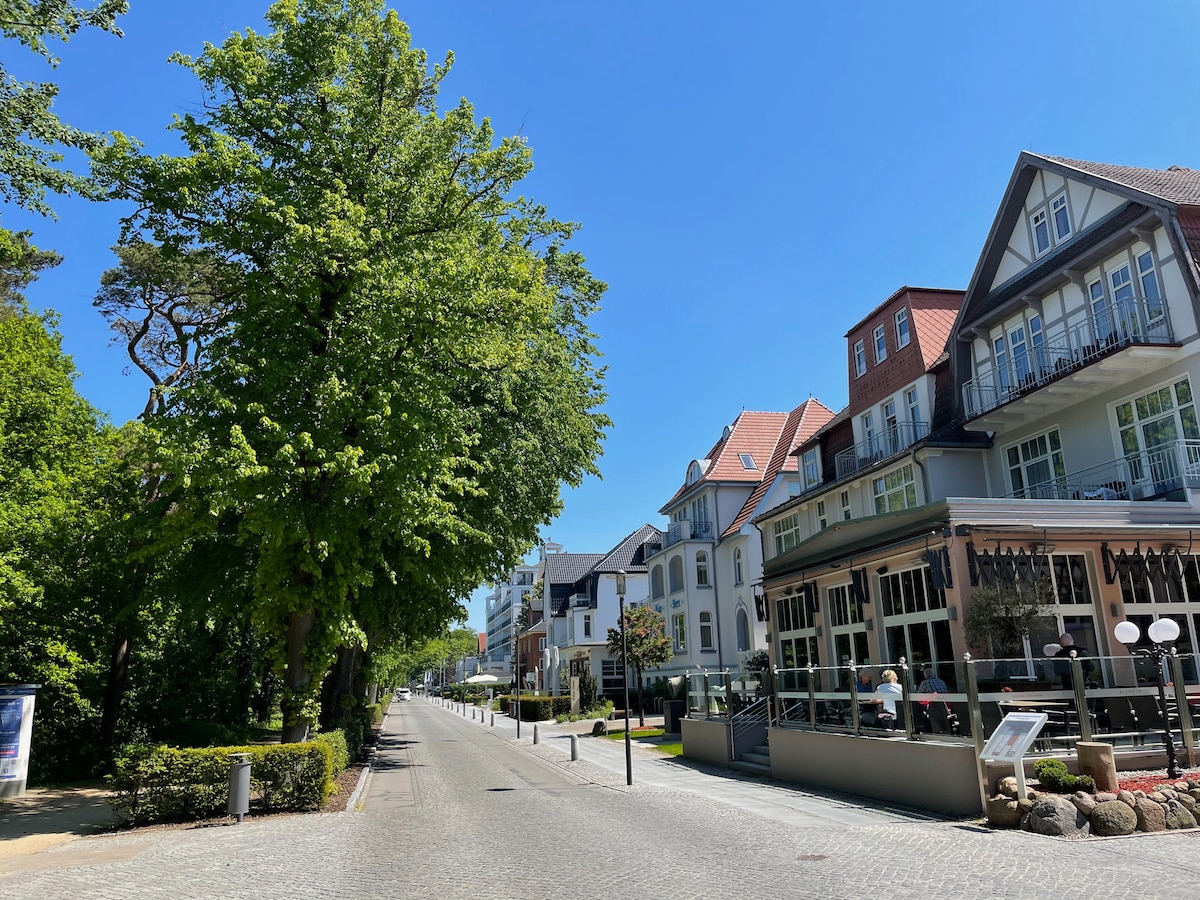 A broad street is lined with tall trees and charming buildings. Several structures feature balconies and outdoor seating areas, with a café visible on the right. Cobblestone paths are present, highlighting the peaceful ambiance of the area on a clear sunny day.
