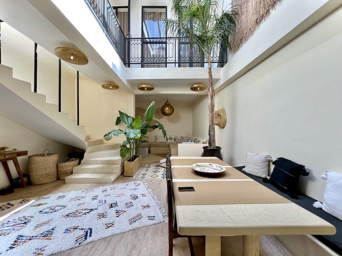 A light-filled patio is depicted, with a spacious dining table positioned centrally. Soft textures and earth-toned decor create a welcoming ambiance. A staircase leads to the upper level, while potted plants add a natural touch. Woven baskets and decorative items enhance the inviting setting.