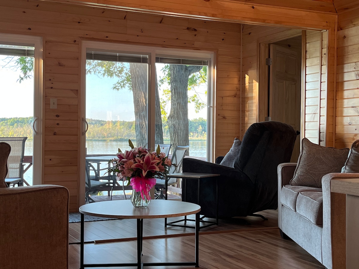 A spacious living area is highlighted by large glass doors opening to a lakeview. A comfortable recliner and a cozy sofa are positioned near a coffee table. Natural light fills the room, showcasing the warm knotty pine walls and inviting atmosphere.
