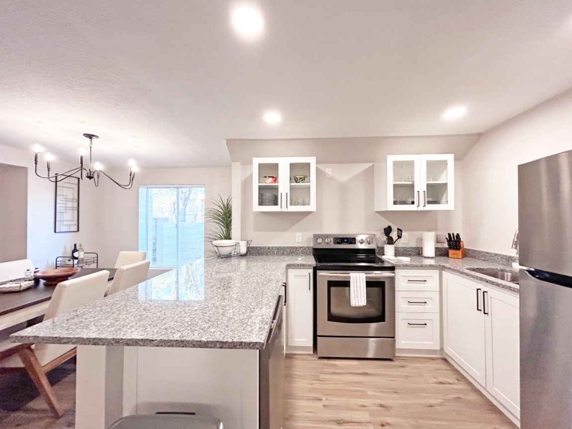 A modern kitchen is presented with sleek granite countertops and stainless steel appliances. White cabinetry provides ample storage, while a dining area is visible in the background. Natural light enters through sliding glass doors, enhancing the spacious and functional layout.