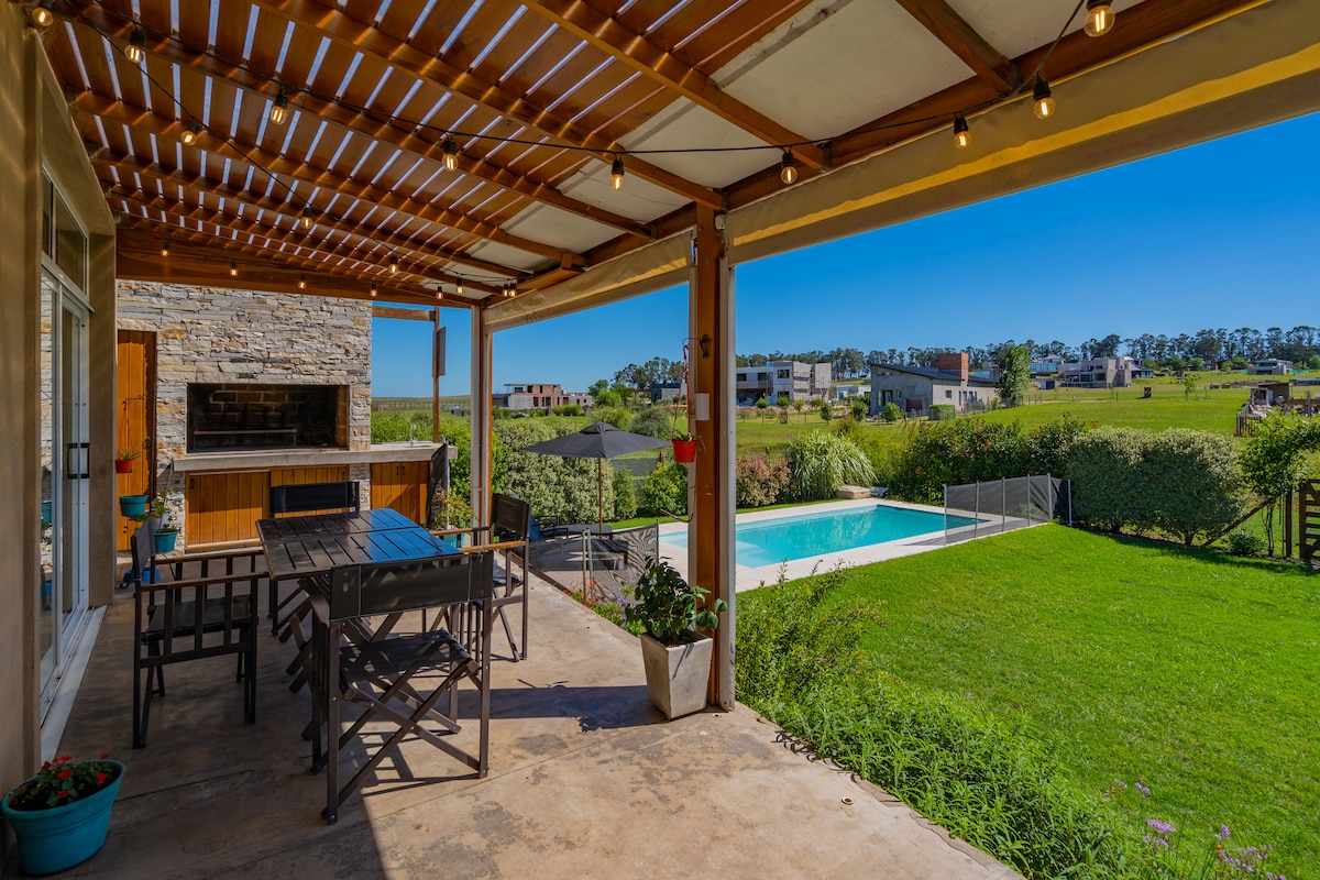 An outdoor patio area is presented with a wooden pergola overhead, accented by string lights. A large dining table is set for gatherings, with a view of the private swimming pool and lush green lawn extending into the natural landscape.