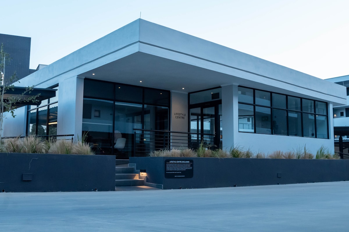 The exterior of a modern building is showcased, featuring large glass windows that allow natural light to filter through. A sleek entrance with steps is visible, complemented by neatly arranged landscaping and soft lighting along the pathway.